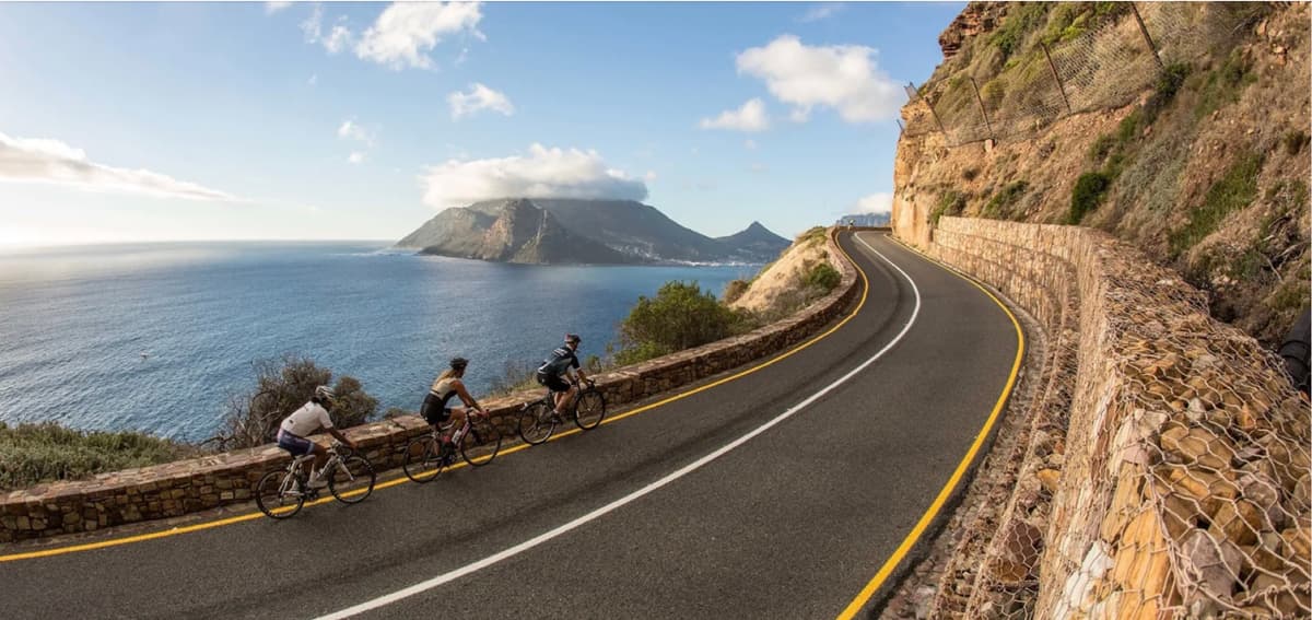 Three road cyclists rounding a curve on Chapman's Peak Drive above the Atlantic, with the Sentinel and the Hout Bay shoreline behind