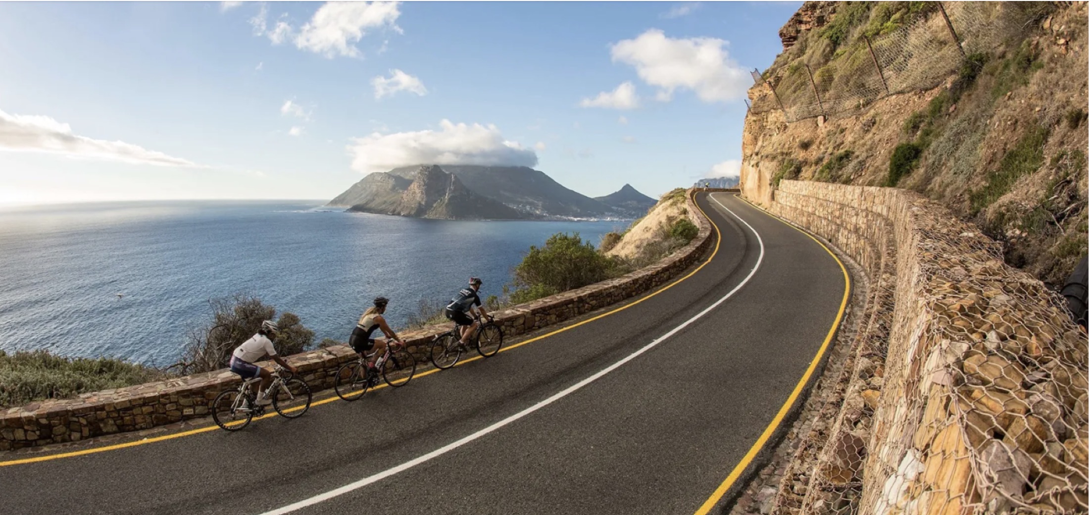 Three road cyclists rounding a curve on Chapman's Peak Drive above the Atlantic, with the Sentinel and the Hout Bay shoreline behind